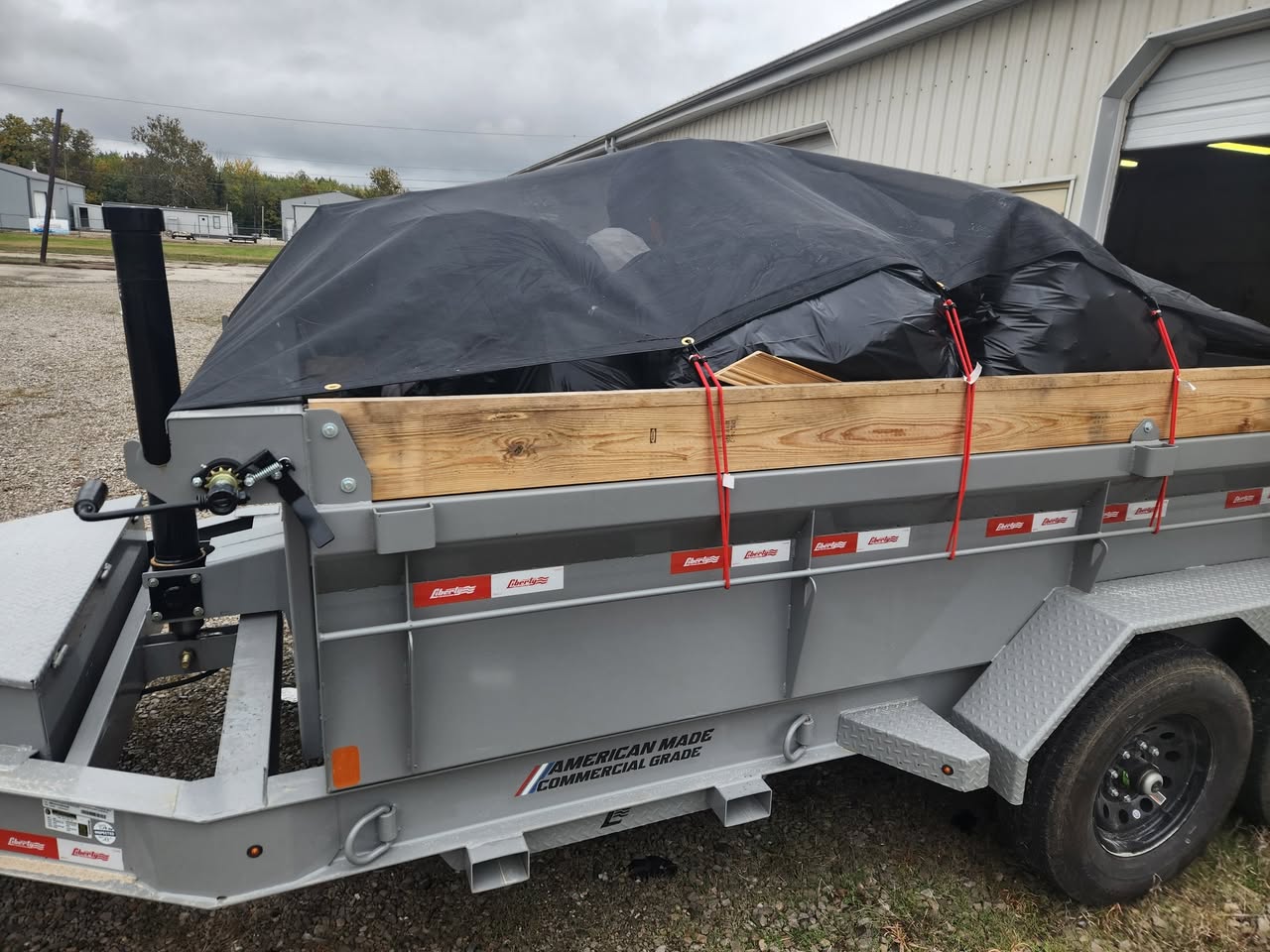 Dump trailer filled with junk and covered with a black tarp, secured with red straps for transport.