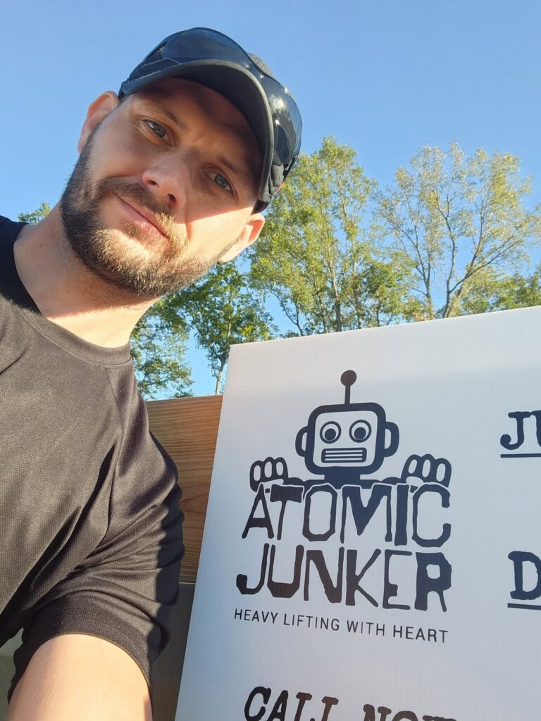Man taking a selfie next to an Atomic Junker sign featuring a robot logo and the tagline ‘Heavy Lifting With Heart,’ promoting the junk removal service.
