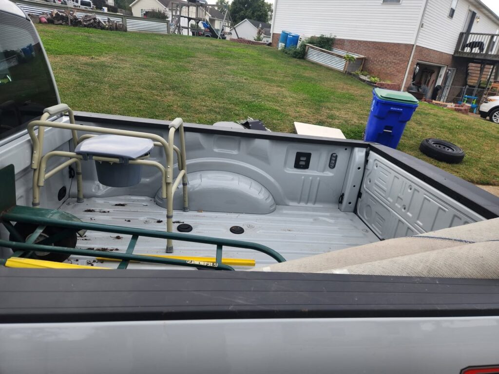 Pickup truck bed loaded with miscellaneous junk items, including a bedside commode and yard equipment.