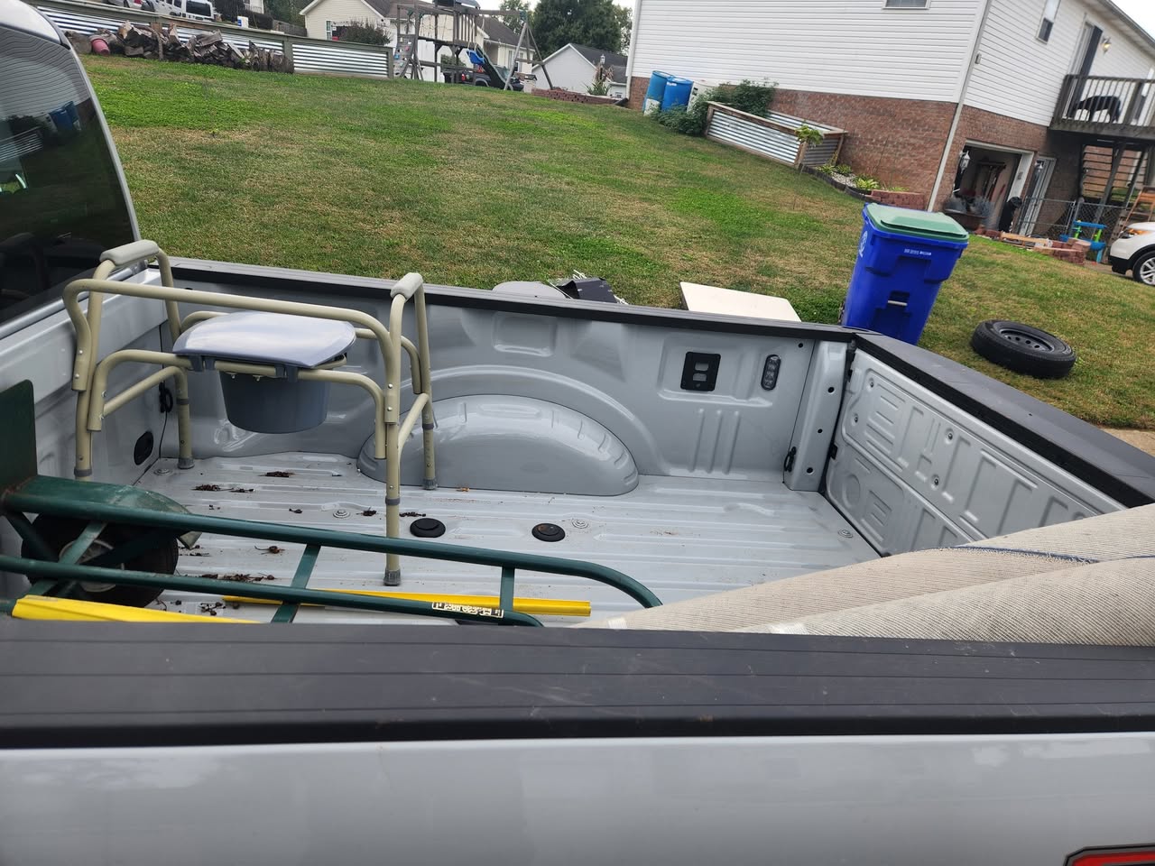 Pickup truck bed loaded with miscellaneous junk items, including a bedside commode and yard equipment.