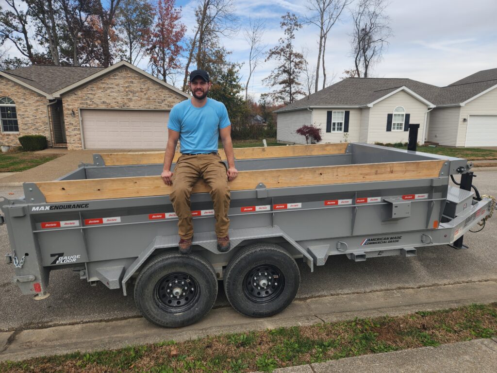Junk removal worker sitting on an empty dump trailer in a suburban driveway, showing the equipment used for debris hauling and cleanout services.